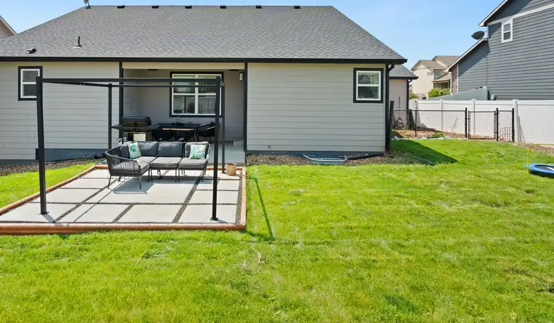 Square backyard slab patio under a black metal pergola behind a new single-story home